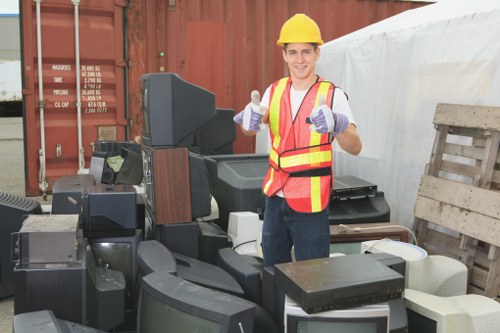 A smiling man wearing a yellow safety helmet, high-visibility vest with orange and yellow stripes, and purple work gloves is standing outdoors at a rubbish removal site. He is giving a thumbs-up gesture and is surrounded by a variety of discarded electronic items and broken appliances, including computer monitors, keyboards, and other damaged electronic devices. In the background, there is a large shipping container with visible weight markings and a weathered wooden pallet leaning against a white covering or tent. The ground surface appears to be a concrete or tarmac area, typical of a waste management or clearance operation in the Hammersmith area. The scene captures a professional rubbish clearance environment, consistent with services provided by Hammersmith House Clearance, with a focus on environmental safety and responsible waste disposal.