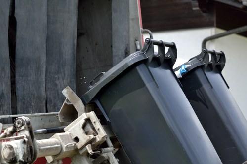 Close-up view of two dark grey plastic wheelie bins positioned side by side on a paved driveway or alleyway. The bins have smooth, slightly reflective surfaces with rounded lids that are partially open. They are secured with black handles that are visible at the top. The background features a section of a wooden fence or paneling, and the bins are attached to a metal frame or trolley, with part of a lifting mechanism visible on the left side of the image. The environment suggests a typical rubbish collection or household waste disposal area, consistent with services offered by Hammersmith House Clearance in the London district. The lighting is neutral, providing clear visibility of the bins’ texture and placement, suitable for a professional waste removal context.