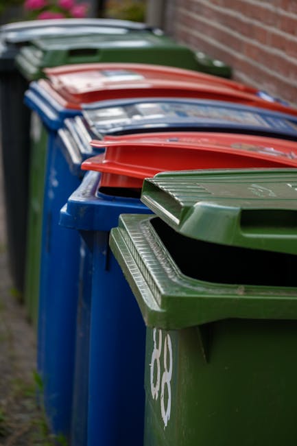 A person's hand is visible gripping the top of a green and blue plastic rubbish bag, which appears to be full and loosely tied at the top with a knotted plastic tie. The bag's surface is shiny and sli